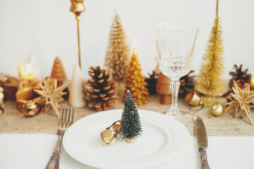 Stylish Christmas table setting. Little christmas tree with bell on plate, vintage cutlery, wineglass, modern golden christmas trees and candles on table. Holiday brunch, new year celebration