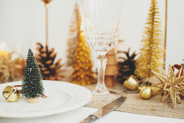 Christmas luxury table setting. Little Christmas tree with bell on plate, vintage cutlery, glasses, golden little christmas trees and ornaments on table. Holiday arrangement of table