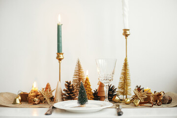 Stylish Christmas table setting. Little christmas tree on plate, vintage cutlery, wineglass, modern golden christmas trees and candles on table. Holiday brunch, new year celebration