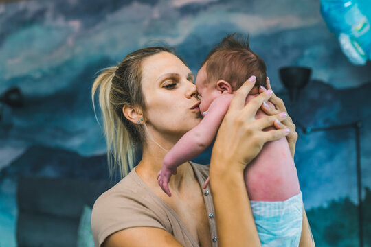 Middle-aged Millennial Caucasian Blonde Mother In Casual Clothing Holding Up Her Little Infant Baby Boy In Diaper And Giving Him A Kiss On His Nose. Closeup Indoor Shot. Blue Wallpaper In The