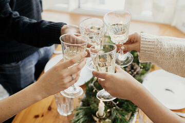 Hands with champagne glasses clinking on background of stylish table setting with fir branches with golden lights and candles. Friends toasting with champagne and celebrating. Christmas feast