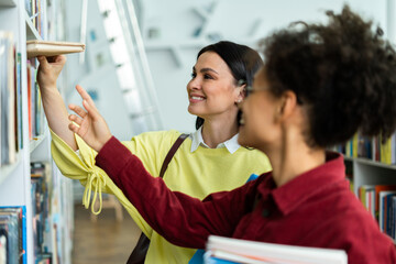 Two student women taking book from library bookshelf. Young librarian searching books and taking one book from library bookshelf