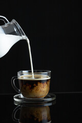 Coffee with cream. Pouring the cream from the pitcher into the coffee. Black background. Copy Space.