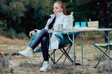 camping, a beautiful woman is sitting at a tourist table in an armchair and enjoying unity with nature in a pine forest