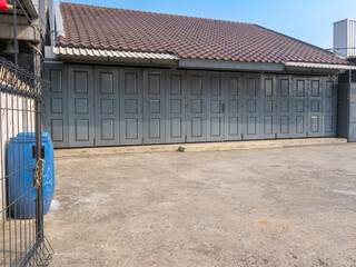 A warehouse area with closed grey doors, a blue garbage bin, an open gate, and a spacious parking area, under a clear blue sky.