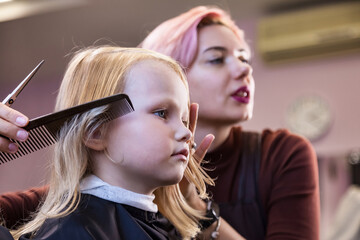 Hairdresser makes hairdo for baby in barber shop. Hair salon, barber woman make fashionable pretty hairstyle for cute little blond girl child in modern barbershop. Hairstyles, beauty concept