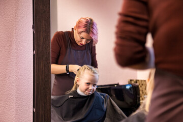 Hairdresser makes hairdo for baby in barber shop at mirror. Hair salon, Barber woman make fashionable braids hairstyle for cute little blond girl child in modern barbershop. Concept hairstyle, beauty