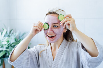 A young smiling woman with pink clay facial mask holds cucumber slices making a refreshing eye mask...