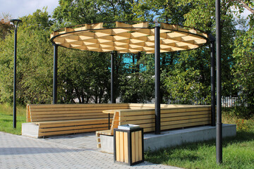 A light wooden gazebo with a bench, a table and an unusual hutch in a city park. Urban architecture