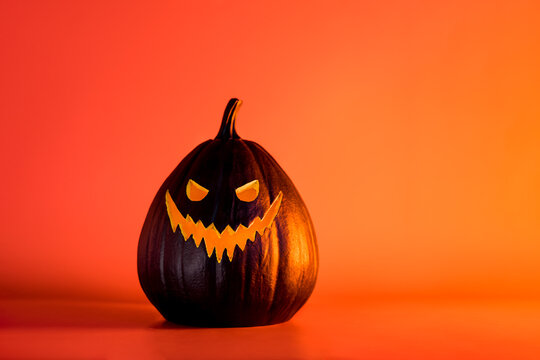 Spooky halloween black pumpkin, Jack O Lantern, with illuminated evil face and eyes on bright orange background with copy space. Happy Halloween decor concept. Festive postcard. Selective focus