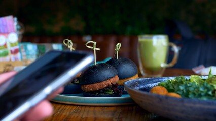 At a restaurant table, a man sits on his phone and looks at the screen by swiping his fingers over it. On the table are salad dishes and three hamburgers with various fillings and black buns.