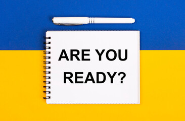 Fototapeta premium A white notebook with the text ARE YOU READY and a white pen on a blue and yellow background.