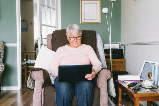 Senior grandmother sitting on armchair, looking at laptop screen. Grey-haired mature woman reading news, chatting with family, watching video, online consulting with doctor. Seniors and technology