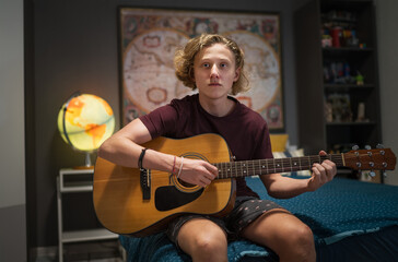 Portrait of teenager boy sitting on the cozy sofa and playing acoustic guitar dressed casual...