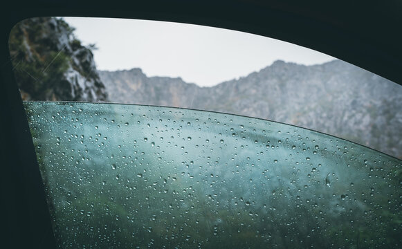 Half opened front car door misted window covered with a cold rain drops with a rocky mountains range and heavy stormy clouds.