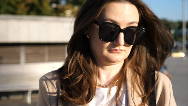 Portrait of woman in sunglasses with displeased facial expression. Serious brunette girl looks into camera at city street. Unhappy irritated businesswoman. Concept of sorrowful or annoyed emotion