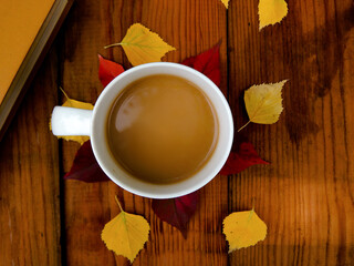 On a wooden table there is a white mug with coffee around the mug lies fallen leaves, next to it lies a book with a brown cover