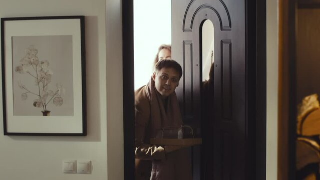 Medium long of Caucasian woman and man quietly entering apartment, husband holding cake, smiling