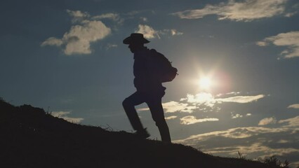 Male traveler in hat enjoys hiking in wild area at back sunset light exploring nature. Dark silhouette of adventurous man with backpack going uphill top against sky with clouds on horizon at twilight