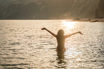 The girl stands in the sea and looks ahead at the sunset. Her hands are raised up. She wears a black swimsuit and her hair is down.