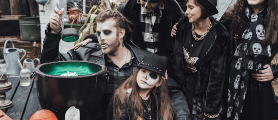 Obraz premium Scary family, mother, father, daughters celebrating halloween. Cauldron of potion.Terrifying black skull half-face makeup,witch costumes,stylish images.Horror,fun at children's party in barn on street