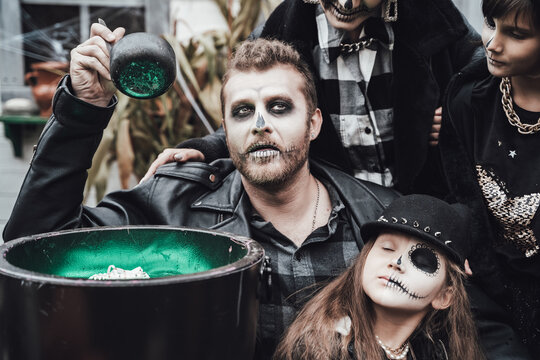 Scary family, mother, father, daughters celebrating halloween. Cauldron of potion.Terrifying black skull half-face makeup,witch costumes,stylish images.Horror,fun at children's party in barn on street