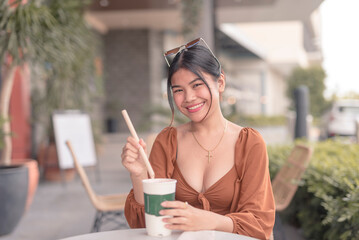 A smiling Southeast Asian woman sitting outside the coffee house places the straw inside her drink.