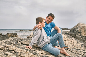 Fototapeta premium Happy family, father and son bonding, sitting on stone by the sea looking at view enjoying summer vacation. Togetherness Friendly concept