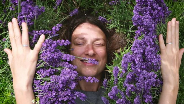 Touching nature. Young happy woman lying among lavander flowers in blossom field, hiding in the flowers. Female without makeup relax on the grass on sunny summer day.
