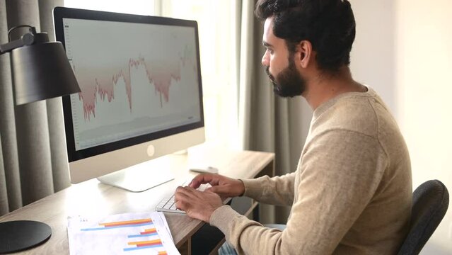 Frelancer man is working on project on the distance, analyzes the market. Determined young bearded Indian trader worker in casual clothes sitting at the desk
