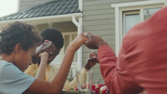 Waist up of African American family holding hands and praying while having lunch together outdoors at backyard on summer day