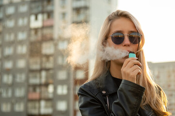 Young woman with sunglasses smoke vape pod system on a city street against the background of buildings. Female inhales and exhales vapor of electronic cigarette.