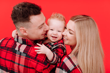 smiling young mother and father are kissing and hugging their baby boy on red background. concept...