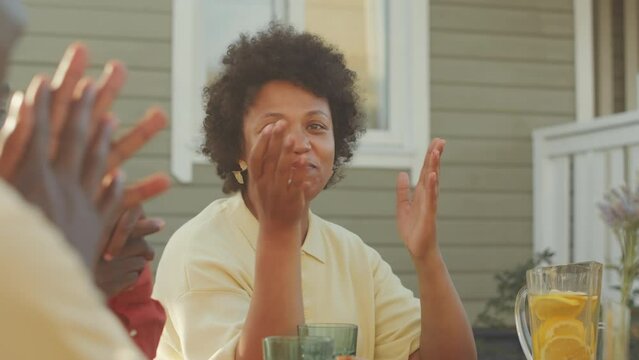 Waist up of smiling Black woman praying at dinner table while getting together outdoors in yard with family