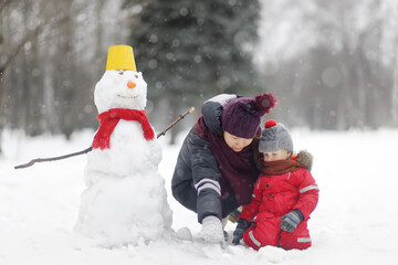 Fototapeta na wymiar Little child with young nanny playing with snowman in park. Active winter leisure with kids.