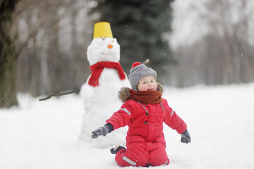 Fototapeta na wymiar Little boy in red clothes having fun with big snowman. Child during stroll in a snowy winter park. Active winter outdoor leisure for children.