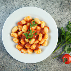 Gnocchi in sugo sauce with some parsley served in white plate on burnt cement table with pieces of tomato and parsley scattered on the table, Top view