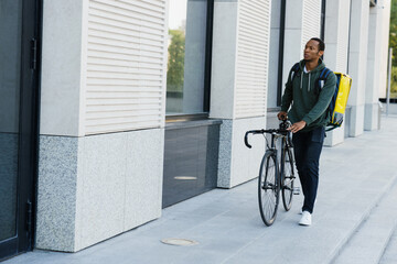 A dark-skinned male courier walks with a bicycle along the building. Delivery of dishes from the restaurant.
