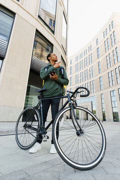 Delivery in the city by bike. An African American male courier with a yellow backpack holds a mobile phone and looks at the delivery address.