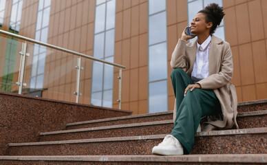 Fototapeta premium african american young woman talking on the phone while sitting on the steps against the backdrop of a business center