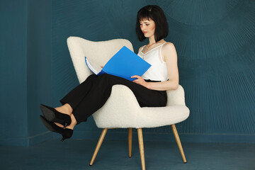 Young self confident woman with short hair sitting, relaxing on chair and holding blue folder and...