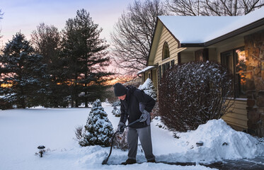 Senior man shoveling driveway in front of his house after snowfall in Midwest at sunset