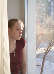 A teenage girl sits on the windowsill and looks thoughtfully out the window at the snow-covered street. Banner, flyer, layout design, postcard. Russia. Omsk.
