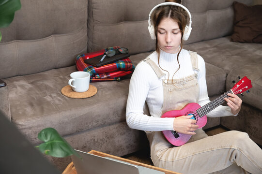 Young caucasian hipster woman is engaged in online ukulele lesson at home, sitting on the floor, on the background of the sofa, in the living room