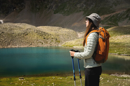 a young caucasian woman with a backpack and in a panama, stands with trekking poles near a mountain lake and looks into the distance. hiking, walking