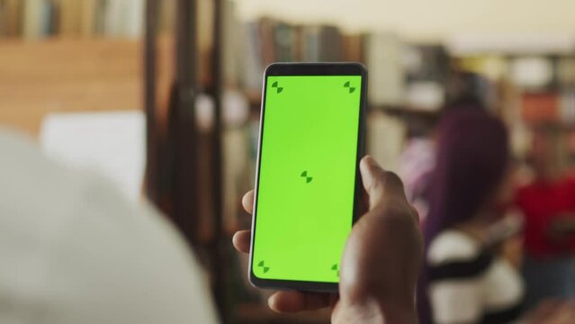 Closeup of a person in a white shirt holding smart phone device with a green screen chromakey vertically in their hand, looking at app, at the indoor public space, library or a bookstore