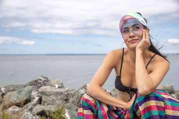Asian woman wearing a visor and bikini top with colorful striped pants rests her hand under her chin and looks up while thinking. Ocean water is behind her as she is sitting on shoreline rocks. 