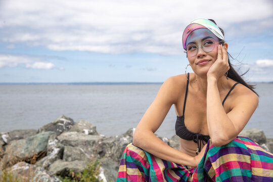 Asian woman wearing a visor and bikini top with colorful striped pants rests her hand under her chin and looks up while thinking. Ocean water is behind her as she is sitting on shoreline rocks. 