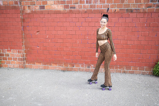 Asian woman wearing a two piece, checkered body suit and roller skates smiling next to a brick wall. 
