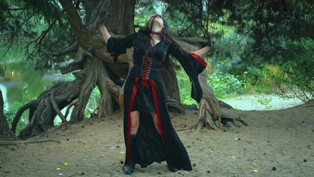 Fantasy portrait woman witch dancing a ritual dance. Sexy girl magician shakes her head hand, long hair fly in wind slow motion. Black red carnival dress costume. Tree roots green forest water river